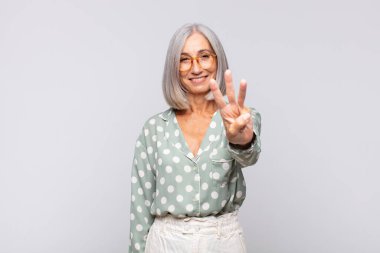 Gri saçlı kadın gülümsüyor ve arkadaş canlısı görünüyor, üç ya da üç numarayı gösteriyor, eli önde, geriye doğru sayıyor.