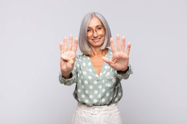 Gri saçlı kadın gülümsüyor ve arkadaş canlısı görünüyor. 9 ya da 9 numarayı gösteriyor. Eli önde, geriye doğru sayıyor.