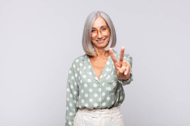 Gri saçlı kadın gülümsüyor ve arkadaş canlısı görünüyor. İki ya da iki numarayı gösteriyor. Elleri önde, geri sayıyor.