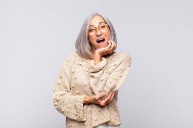 Gri saçlı kadın şok ve inançsızlıkla ağzı açık, elleri yanağında ve kolları çapraz, sersemlemiş ve şaşırmış hissediyor.