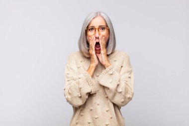 Gri saçlı kadın şok olmuş ve korkmuş hissediyor, ağzı açık ve elleri yanaklarında korkmuş görünüyor.
