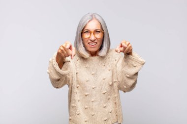 Gri saçlı kadın kamerayı işaret ederek hem parmaklarını hem de kızgın yüzünü göstererek sana görevini yapmanı söylüyor.