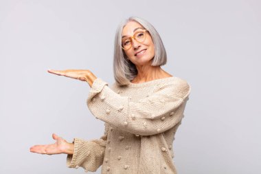 Gri saçlı kadın bir objeyi iki eliyle fotokopi alanında tutuyor, bir objeyi gösteriyor, sunuyor ya da reklamını yapıyor.