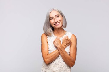 Gri saçlı kadın neşeli bir şekilde gülümsüyor ve kutluyor, yumruklarını sıkıp kollarını kavuşturuyor, mutlu ve pozitif hissediyor.