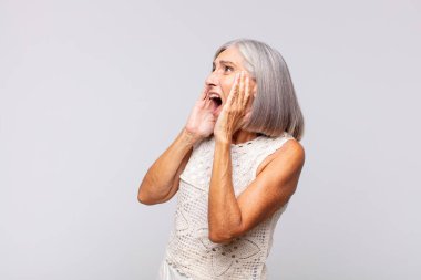 Gri saçlı kadın mutlu, heyecanlı ve şaşırmış hissediyor, iki eli yüzü yana bakıyor.