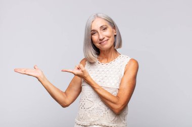 Gri saçlı kadın neşeyle gülümsüyor ve yan taraftaki fotokopi alanına işaret ediyor, bir nesneyi gösteriyor veya reklamını yapıyor.