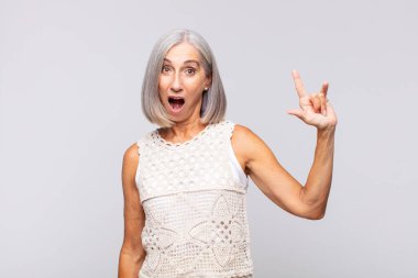 Gri saçlı kadın mutlu, eğlenceli, kendinden emin, pozitif ve asi hissediyor, elle kaya veya heavy metal işareti yapıyor.