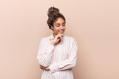 Genç afro bir kadın mutlu, kendinden emin bir ifadeyle gülümsüyor, elleri çenesinde, merak ediyor ve yan tarafa bakıyor.