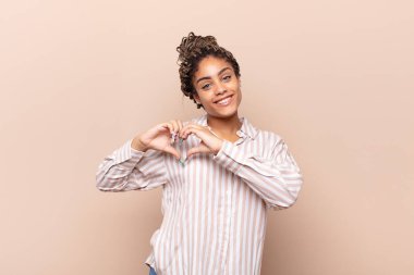 Genç afro kadın gülümsüyor ve mutlu, sevimli, romantik ve aşık, iki eliyle kalbi şekillendiriyor.