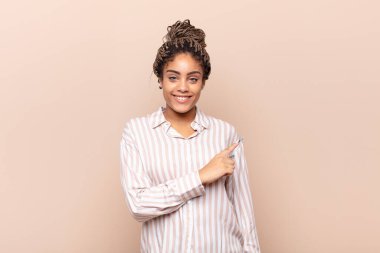 Genç afro kadın neşeyle gülümsüyor, mutlu hissediyor ve yan ve yukarı doğru işaret ediyor, fotokopi alanında nesneler gösteriyor.