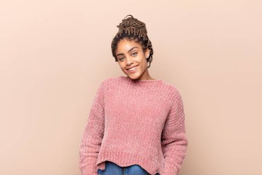 Genç afro kadın neşeli ve tesadüfen gülümsüyor, olumlu, mutlu ve kendinden emin bir bakışla göğüs göğüse tutuşuyor.