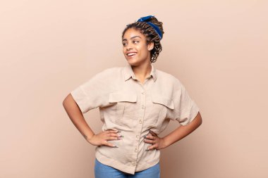 Genç afro kadın mutlu, neşeli ve kendinden emin görünüyor, gururla gülümsüyor ve iki elini de kalçasında istiyor.