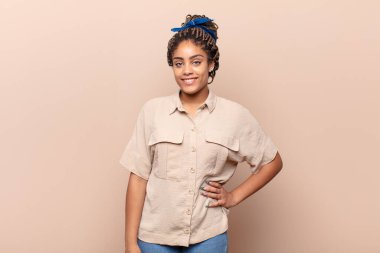 Genç afro kadın mutlu bir şekilde gülümsüyor, eli kalçasında ve kendinden emin, pozitif, gururlu ve arkadaş canlısı.