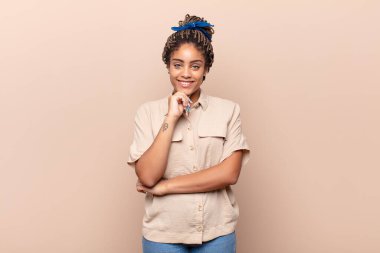 Genç afro kadın mutlu görünüyor ve gülümsüyor, eli çenesinde, merak ediyor ya da soru soruyor, seçenekleri karşılaştırıyor.