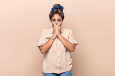 Genç afro kadın mutlu, neşeli, şanslı ve şaşkın görünüyor. İki eliyle ağzını kapatıyor.