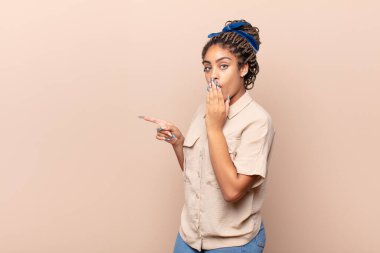 Genç afro kadın kendini mutlu, şaşkın ve şaşırmış hissediyor. Ağzını eliyle kapatıyor ve yanal fotokopi alanını işaret ediyor.