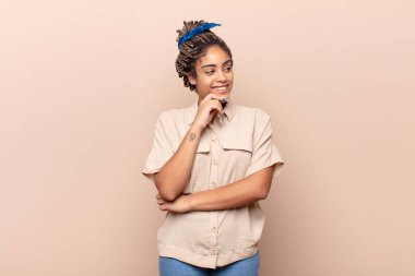 Genç afro bir kadın mutlu, kendinden emin bir ifadeyle gülümsüyor, elleri çenesinde, merak ediyor ve yan tarafa bakıyor.