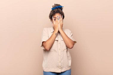 Genç afro kadın elleriyle yüzünü kapatıyor, parmaklarının arasından şaşkın bir ifadeyle yan tarafa bakıyor.
