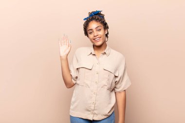 Genç afro kadın mutlu ve neşeyle gülümsüyor, el sallıyor, seni selamlıyor ve selamlıyor, ya da hoşça kal diyor.