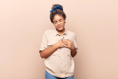 Genç afro kadın üzgün, yaralı ve kalbi kırık görünüyor, iki elini de kalbe yakın tutuyor, ağlıyor ve bunalımda.
