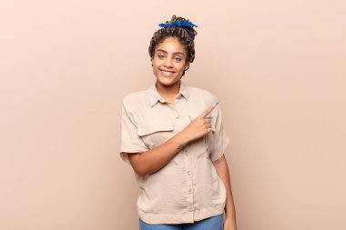 Genç afro kadın neşeyle gülümsüyor, mutlu hissediyor ve yan ve yukarı doğru işaret ediyor, fotokopi alanında nesneler gösteriyor.