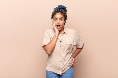 Genç afro kadın şok ve inançsızlık içinde, elleri yanak ve kolları çapraz, sersemlemiş ve şaşırmış hissediyor.
