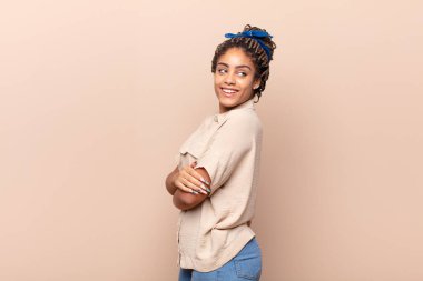 Genç afro kadın neşeyle gülümsüyor, mutlu, tatmin olmuş ve rahatlamış hissediyor, kollarını kavuşturup yan tarafa bakıyor.