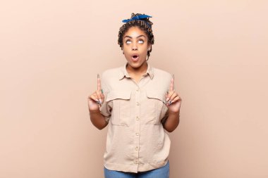 Genç afro kadın şaşırmış, şaşırmış ve ağzı açık görünüyor, iki eliyle uzayı işaret ediyor.