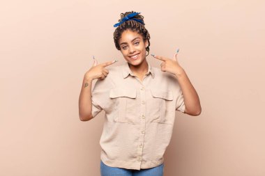 Genç afro kadın kendine güvenerek gülümsüyor, kendi geniş gülüşünü gösteriyor, pozitif, rahat, tatmin olmuş tavır.