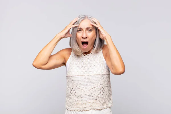 stock image gray haired woman feeling horrified and shocked, raising hands to head and panicking at a mistake