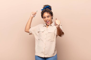 Genç afro kadın muzaffer bir şekilde bağırıyor, heyecanlı, mutlu ve şaşırmış görünüyor, kutlama yapıyor.