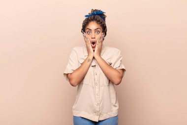 Genç afro kadın şok olmuş ve korkmuş hissediyor, ağzı açık ve elleri yanaklarında korkmuş görünüyor.
