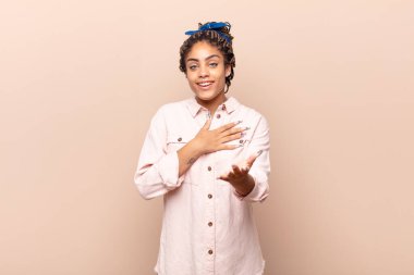 Genç afro kadın mutlu ve aşık, bir eli kalbinin yanında, diğer eli önde uzanmış, gülümsüyor.