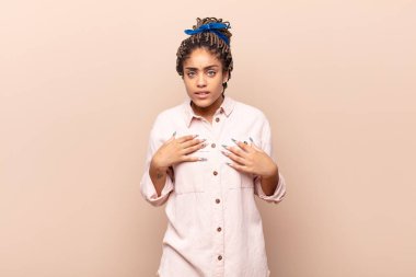 Genç afro kadın şaşkın ve şaşkın bir bakışla kendini işaret ediyor. Seçildiğine şaşırmış ve şaşırmış.