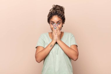 Genç afro kadın mutlu, neşeli, şanslı ve şaşkın görünüyor. İki eliyle ağzını kapatıyor.