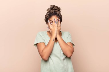 Genç afro kadın elleriyle yüzünü kapatıyor, parmaklarının arasından şaşkın bir ifadeyle yan tarafa bakıyor.