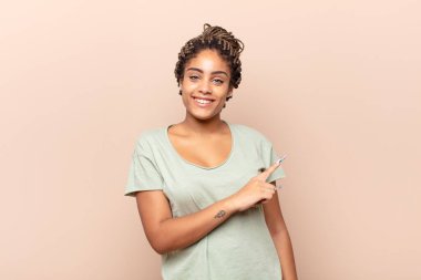 Genç afro kadın neşeyle gülümsüyor, mutlu hissediyor ve yan ve yukarı doğru işaret ediyor, fotokopi alanında nesneler gösteriyor.