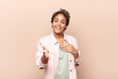 Genç afro kadın mutlu ve aşık, bir eli kalbinin yanında, diğer eli önde uzanmış, gülümsüyor.