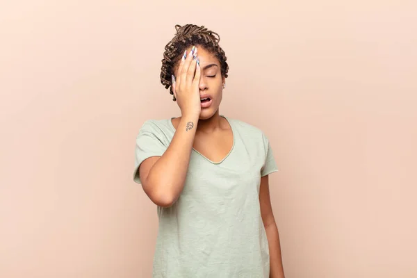 young afro woman looking sleepy, bored and yawning, with a headache and one hand covering half the face