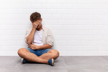 Genç adam stresli, utanmış ya da üzgün görünüyor, baş ağrısı var, yüzü elle kapatılmış.