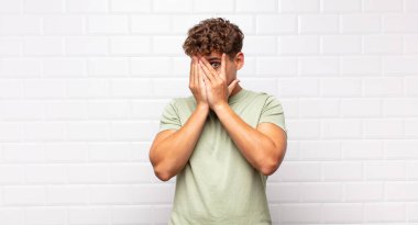 Genç adam elleriyle yüzünü kapatıyor, parmaklarının arasından şaşkın bir ifadeyle yan tarafa bakıyor.
