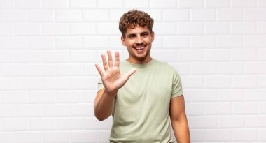 Genç adam gülümsüyor ve arkadaş canlısı görünüyor, 5 ya da 5 numarayı gösteriyor, eli önde, geriye doğru sayıyor.