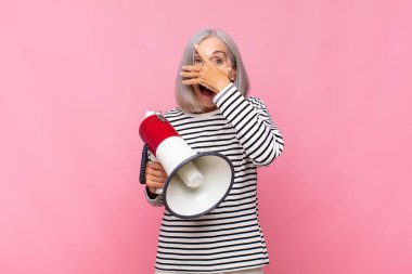 Orta yaşlı bir kadın şok olmuş, korkmuş ya da korkmuş görünüyor. Yüzü elle örtülmüş ve megafonla parmaklarının arasından bakıyor.