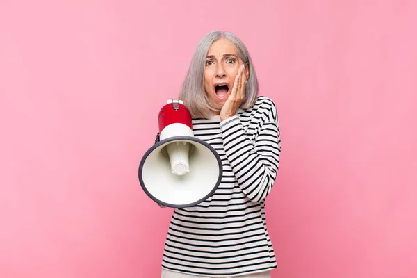 middle age woman feeling shocked and scared, looking terrified with open mouth and hands on cheeks with a megaphone