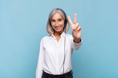 Orta yaşlı bir kadın gülümsüyor ve mutlu görünüyor, kaygısız ve pozitif, tek eliyle zafer ya da barış işareti yapıyor. tele pazarlamacı kavramı