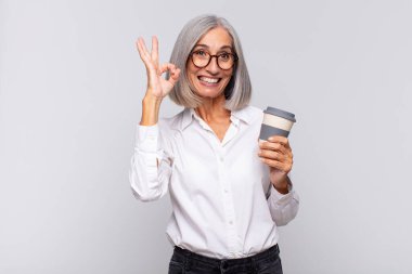 Orta yaşlı bir kadın mutlu, rahat ve tatmin olmuş hissediyor. Onayını iyi bir jestle, gülümseyen bir kahve konseptiyle gösteriyor.