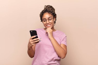 Genç afro bir kadın mutlu, kendinden emin bir ifadeyle gülümsüyor, çenesi elleri çenesinde, merak ediyor ve yan tarafa bakıyor. akıllı telefon kavramı