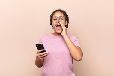 Genç afro kadın mutlu, heyecanlı ve pozitif hissediyor, elleri ağızlarının yanında bağırarak bağırıyor. akıllı telefon kavramı