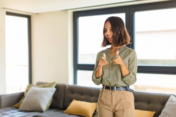 young latin woman feeling happy, cool, satisfied, relaxed and successful, pointing at camera, choosing you. home concept