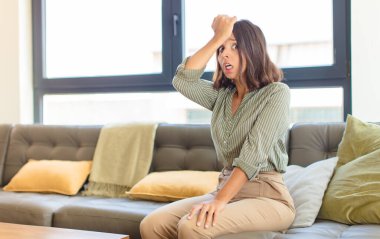 young latin woman raising palm to forehead thinking oops, after making a stupid mistake or remembering, feeling dumb. home concept